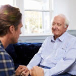 Adult son holding hands with his smiling senior father on a sofa at home
