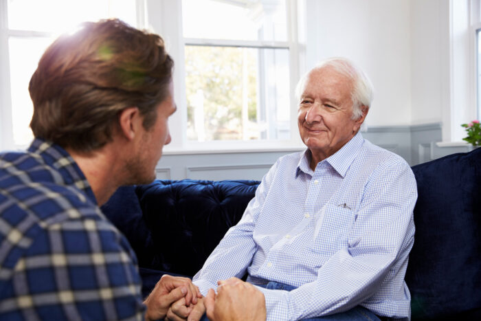 Adult son holding hands with his smiling senior father on a sofa at home