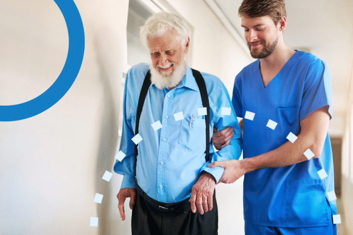 Caregiver assisting elderly man walking in a senior living facility hallway.