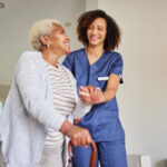 A young caregiver in blue scrubs smiles warmly while assisting an elderly woman with a cane. They share a joyful moment indoors, bathed in natural light.