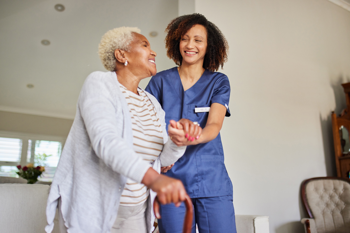 A young caregiver in blue scrubs smiles warmly while assisting an elderly woman with a cane. They share a joyful moment indoors, bathed in natural light.