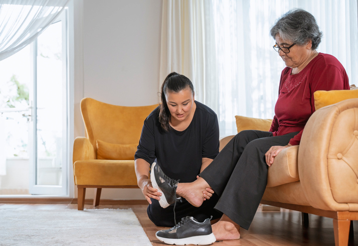 A caregiver kneels on the floor to help an elderly woman put on a sneaker while she sits comfortably on a tan tufted sofa in a bright, warmly furnished living room.