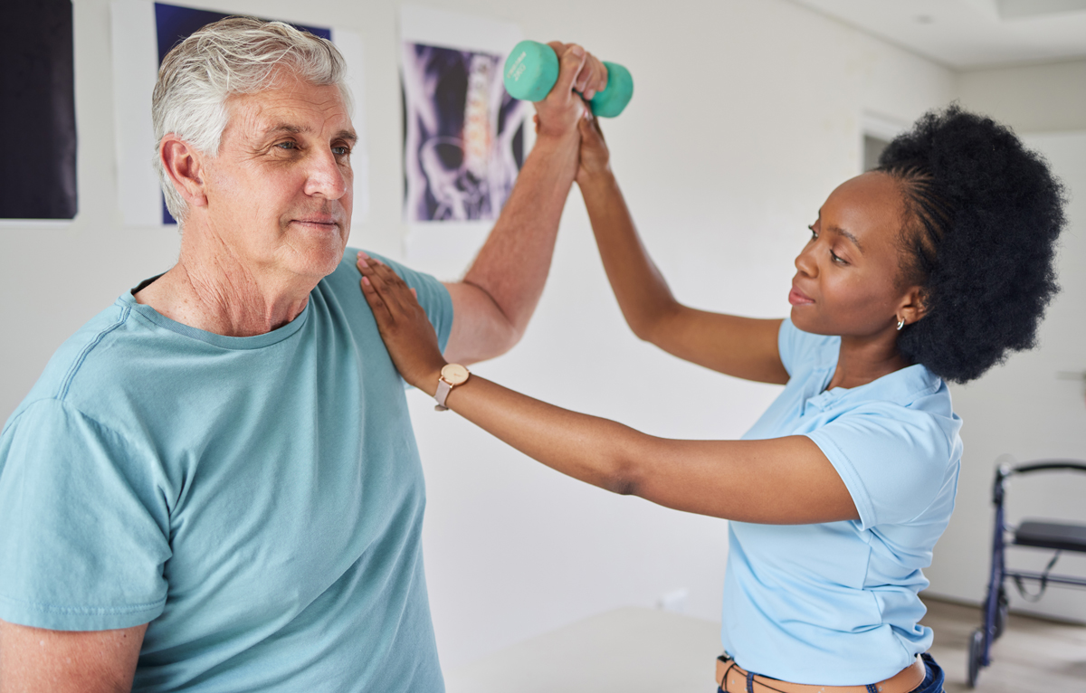 A physical therapist steadies an older man's shoulder while guiding his arm as he raises a teal dumbbell overhead during a rehabilitation session, with X-ray images and a walker visible in the background.